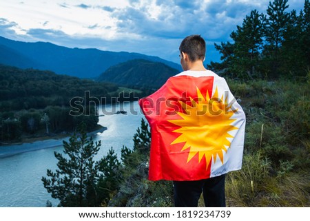 A young man holds the national yazidi flag in his hands against the background of high mountains, forests and rivers