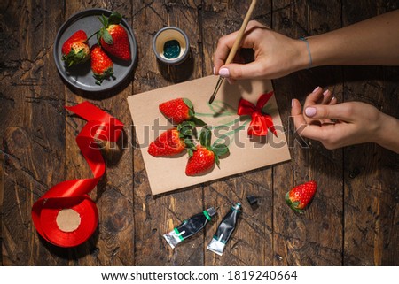 Gift card with live strawberries. A brush in female hands. Girl creates a postcard
