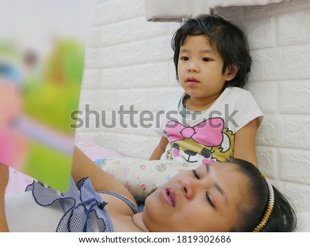 Little Asian baby girl, 3 years old, enjoys listening to her mother reading a bedtime story tale from a book 