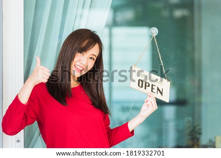 Asian young woman smile show finger thumb up for good sign she notice sign wood board label "WELCOME OPEN" hang through glass door front shop, Business turning open after coronavirus pandemic disease
