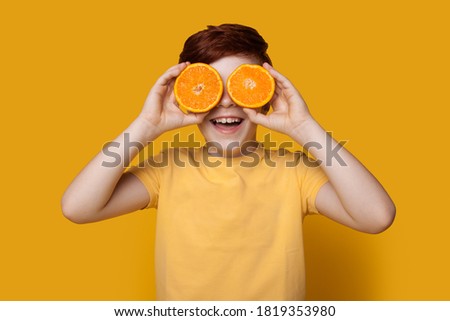 Cute ginger caucasian kid covering eye with sliced oranges posing in a t-shirt on a yellow studio wall Royalty-Free Stock Photo #1819353980