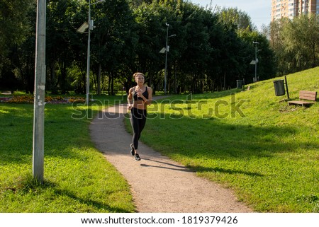 Fit mid-age caucasian woman in sports clothing runs on sunny summer morning in green city park. Sport background. Healthy lifestyle theme. Little motion blur.