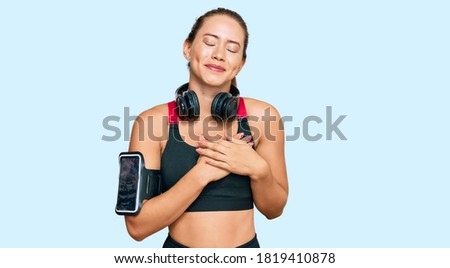 Beautiful blonde woman wearing gym clothes and using headphones smiling with hands on chest with closed eyes and grateful gesture on face. health concept. 