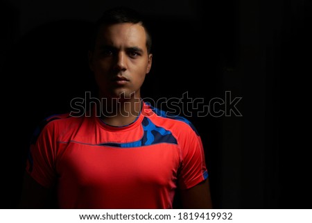 muscular young guy looking forward. Muscular teenage boy wearing orange sport shirt and black background. Young brunette man on black background. Sportsman with short, hanging hair.