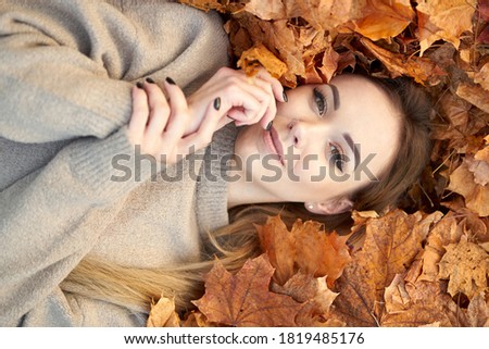 Pretty young girl with charming smile liying in the autumn leaves and enjoying it, holding face and smiling. Top view.                                
