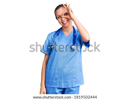 Young beautiful blonde woman wearing doctor uniform and stethoscope doing ok gesture with hand smiling, eye looking through fingers with happy face. 