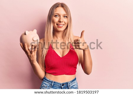 Young blonde woman holding piggy bank smiling happy and positive, thumb up doing excellent and approval sign 