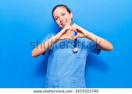 Young blonde woman wearing doctor uniform and stethoscope smiling in love showing heart symbol and shape with hands. romantic concept. 