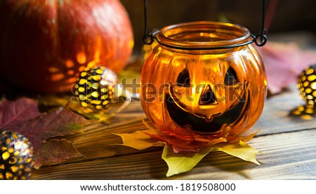 Lamp pumpkin with eyes and mouth made of glass and natural orange pumpkin on a wooden table with yellow and red maple leaves. Halloween, warm autumn atmosphere. Round garland, candle in a candlestick.