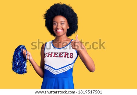 Young african american woman wearing cheerleader uniform holding pompom doing happy thumbs up gesture with hand. approving expression looking at the camera showing success. 