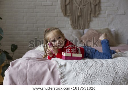 Little cute girl in a red christmas 
sweater  plays on the bed with gifts