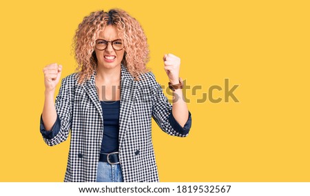 Young blonde woman with curly hair wearing business jacket and glasses angry and mad raising fists frustrated and furious while shouting with anger. rage and aggressive concept.  Royalty-Free Stock Photo #1819532567