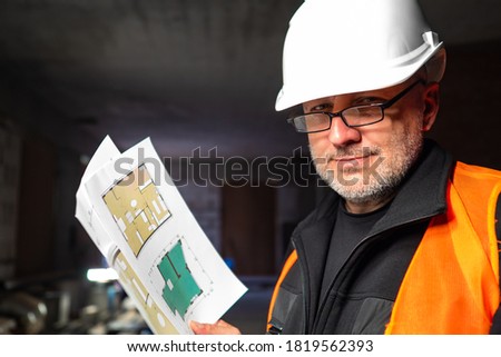 Builder with architectural drawings. Close-up portrait of builder. Man in white helmet looks into camera. Repairman background of building under construction. Architectural plans in hands repairman.