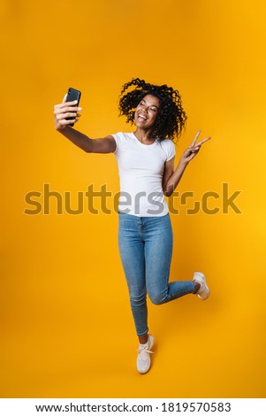 Image of african american woman gesturing peace sign while taking selfie on cellphone isolated over yellow wall