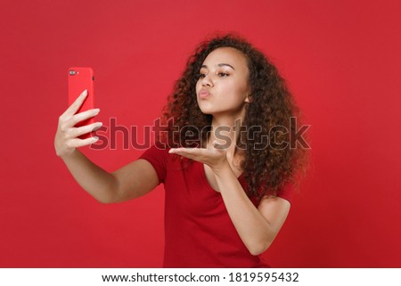 Pretty young african american woman in casual t-shirt posing doing selfie shot on mobile phone blowing sending air kiss isolated on red background studio. People lifestyle concept. Mock up copy space.
