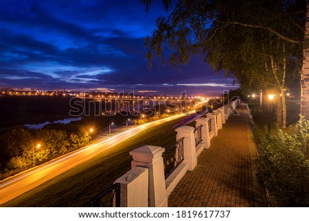 Moving car with blur light through city at night. Bridge over the river and the road. A view from the park from a height with a fence in the foreground. Cityscape.