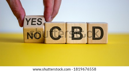 Male hand turns a cube and changes the words 'CBD yes' to 'CBD no' on wooden cubes. Beautiful yellow table, white background, copy space. Conceptual image of CBD legalization and use.