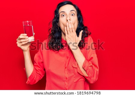 Young beautiful hispanic woman drinking glass of water covering mouth with hand, shocked and afraid for mistake. surprised expression 