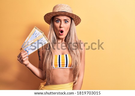Young beautiful tourist woman wearing bikini and hat holding airline boarding pass scared and amazed with open mouth for surprise, disbelief face