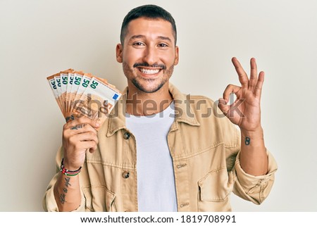 Handsome man with tattoos holding 50 euro banknotes doing ok sign with fingers, smiling friendly gesturing excellent symbol 