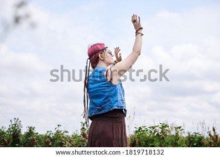 Young hippie woman with short red hair, wearing boho style clothes and sunglasses, dancing on green currant field, posing for picture. Female portrait on natural background. Eco tourism concept.