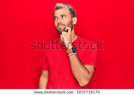 Young handsome blond man wearing casual red t-shirt standing over isolated red background thinking concentrated about doubt with finger on chin and looking up wondering