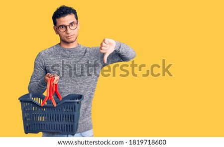 Hispanic handsome young man holding supermarket shopping basket with angry face, negative sign showing dislike with thumbs down, rejection concept 
