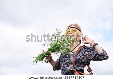 Young blond hippie woman, wearing grey boho style dress and yellow sunglasses, standing on green field, holding camomile bouquet, posing for picture on sunny summer day. Inner freedom concept.
