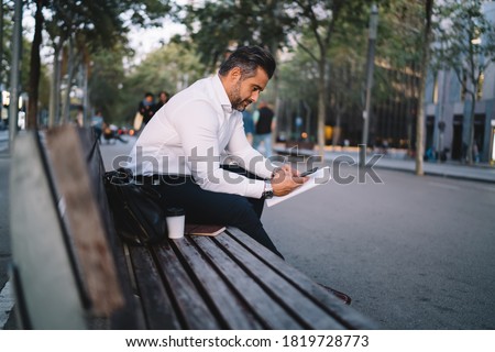 Professional businessman using cellphone gadget for messaging with partner connecting to 4g wireless in downtown, confident male employee with documents sitting at urban bench and making banking