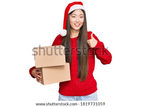 Young chinese woman wearing christmas hat holding delivery package smiling happy and positive, thumb up doing excellent and approval sign 