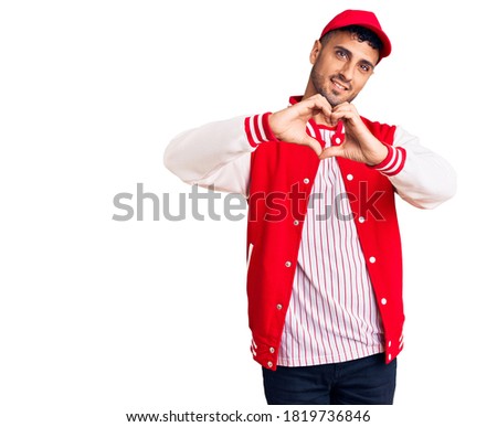 Young hispanic man wearing baseball uniform smiling in love doing heart symbol shape with hands. romantic concept. 