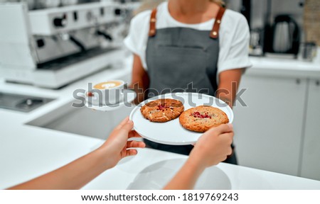 Beautiful female barista giving a cup of coffee or cappuccino and a plate of cookies to a customer in a coffee shop.