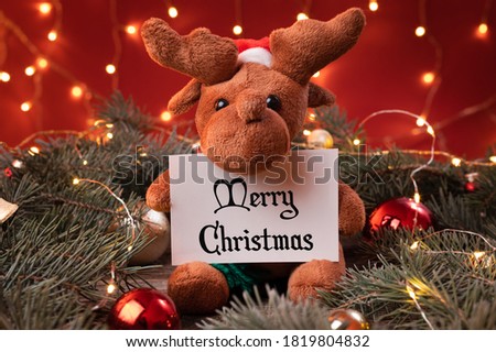 Cute reindeer toy in santa hat holding a white card, sheet of paper with Merry Chrismas in a Christmas scenario.