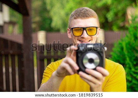 Young happy hipster guy in yellow T-shirt and trendy sunglasses have fun outdoors, takes photo with instant camera on green nature background, resting time. Summer, leisure, photography hobby concept