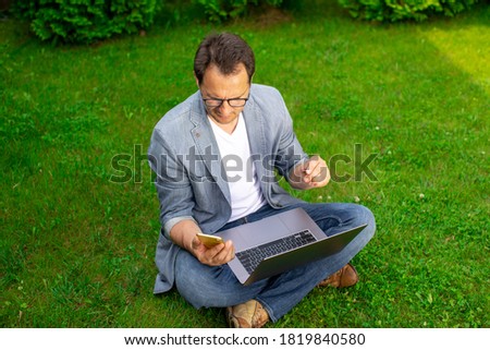 Middle aged businessman works with laptop and mobile phone outdoors, sits on grass in park, sell cryptocurrency, buy up shares, check stock market, online trading, multitasking, busy vacation concept