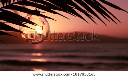 Merry Christmas and Happy New Year greeting card. Christmas decorations glass ball during sunset on palm twig over the sea. Travel to warm countries for New Year holidays concept. Banner, copy space