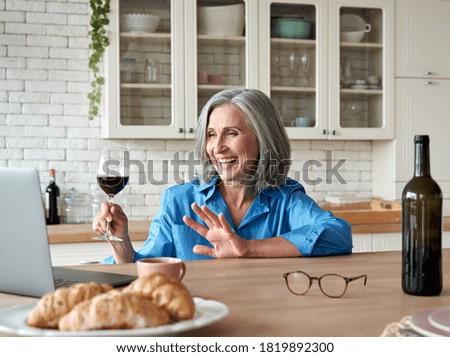 Happy 60s mature woman drinking wine video calling friend on laptop at home. Smiling old middle aged lady holding glass waving hand talking by online social distance chat meeting sit at kitchen table. Royalty-Free Stock Photo #1819892300