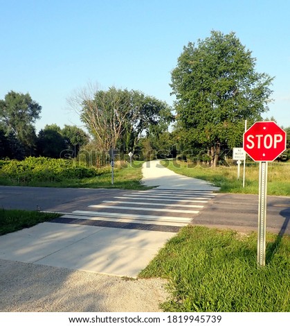 Crossing of a pedestrian and cycling trail with a road. Stop sign for non-motorized users, zebra crossing on the road. No people, rural landscape. Taken in August in south-central Michigan.