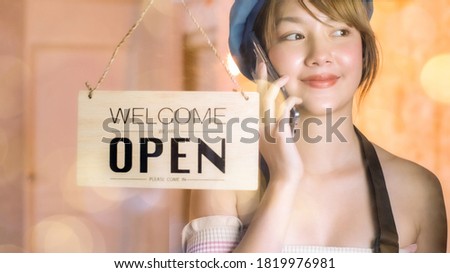 Business owner attractive young Asian woman in apron hanging we're open sign on front door smiling welcoming clients to new cafe. Beautiful Young Cafe Owner Turning Storefront Sign From Close to Open.