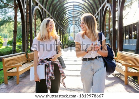 Two happy smiling talking girls teenagers students walking together, young with backpacks, sunny day in the park background
