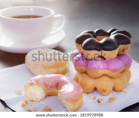 Donuts topped with pink cream that were eaten.  Donuts stacked on top of each other and there is hot coffee behind on dark table