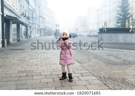 Cute liitle sad lost caucasian girl in warm long pink jacket and hat feel horror on empty street of old european city Wels. Screaming scared alone scared child in town in Morning fog on backround