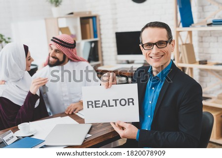 Adult man in glasses holds sign with inscription. Realtor in glasses sits at a desk and holds a sign with the inscription realtor.