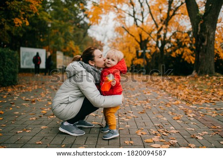 Picture of cheerful family of two. Beautiful caucasian mother in white warm jacket hugs her little child in time their walking in the big park zone.