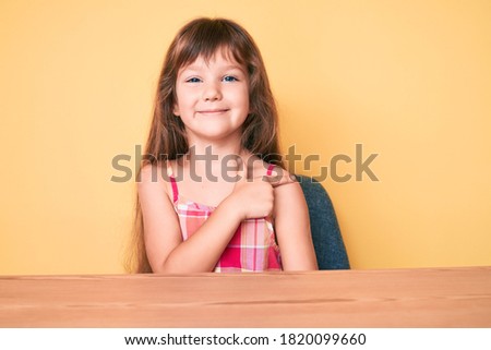 Little caucasian kid girl with long hair wearing casual clothes sitting on the table smiling cheerful pointing with hand and finger up to the side 