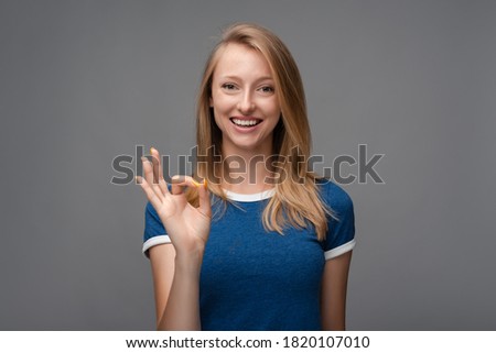 Cheerful girl with blonde straight hair, demonstrating white teeth, with broad smile, looking at the camera with happy expression, showing OK gesture. Human emotions, facial expression concept.