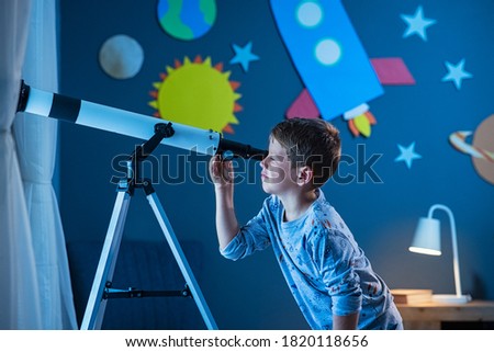 Curious boy using telescope to explore moon surface at night in his bedroom. Young child using telescope to see remote galaxy from room with decorated wall with rocket, planets, stars and spaceship. Royalty-Free Stock Photo #1820118656