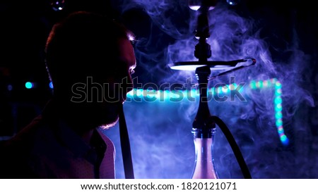 Side view of male silhouette in neon light smoking hookah, relaxing at night club. Media. Man smoking shisha, exhaling thick smoke from his mouth.
