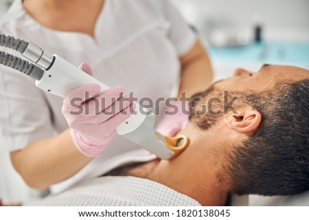 Female esthetician hands in sterile gloves removing unwanted hair from male neck with special laser device Royalty-Free Stock Photo #1820138045