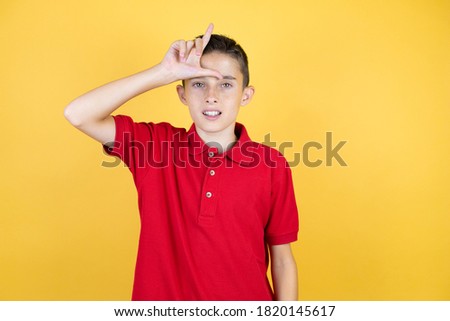 Young beautiful child boy over isolated yellow background making fun of people with fingers on forehead doing loser gesture mocking and insulting.